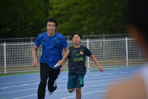 競技場のトラックで、男の子が朝原選手にバトンを渡し、朝原選手が走り出す様子の写真