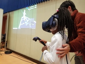 壁に映像が映し出された手前で、女の子がVRフェンシングをつけ、方向などがわかるように付き添われ説明されている様子を左側から写した写真