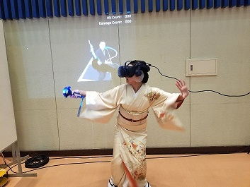 後方の壁に映像が映し出された手前で、着物を着た女性がVRフェンシングを目につけ、左手は手を広げ、膝を曲げVRフェンシングしている様子の写真