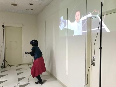 後方の壁にフェンシングしている映像が映り、女性がVRフェンシングをつけ、真正面を向き右手を前に出している写真