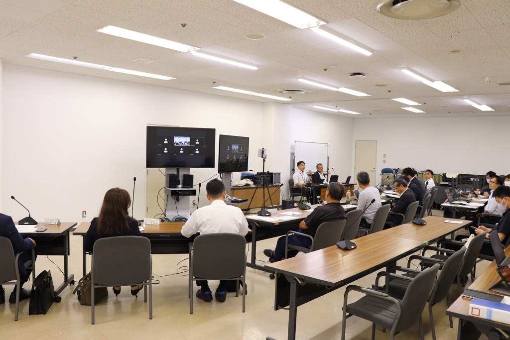 第2回資源循環部会の会議開催中の様子 モニターを軸に半円状に机が並び、委員が座っている。モニターにはZoomでの参加者が映されている。