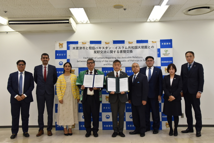 木更津市と中日パキスタン・イスラム共和国大使館との友好交流に関する書簡交換」と日本語で書かれ、下に英語が書かれた横断幕が設置された木更津市のバックボードの前で、書簡を持ったスーツの上に緑色の法被を羽織ったアハマド大使、書簡を持った渡辺市長を中心に、9名の関係者が横一列に並んだ集合写真