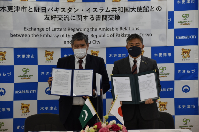 「木更津市と中日パキスタン・イスラム共和国大使館との友好交流に関する書簡交換」と日本語で書かれ、下に英語が書かれた横断幕が設置された木更津市のバックボードの前で、アハマド大使と渡辺市長がフォルダに入った書簡を手に持って並んだ記念写真