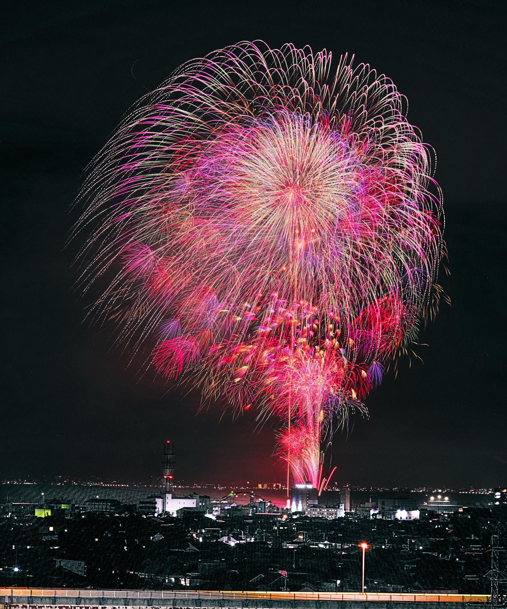 海上に打ちあがった大輪の花火と、その下の3対の打ち上げ花火、噴出花火が色とりどりの光で夜空彩る花火大会の写真