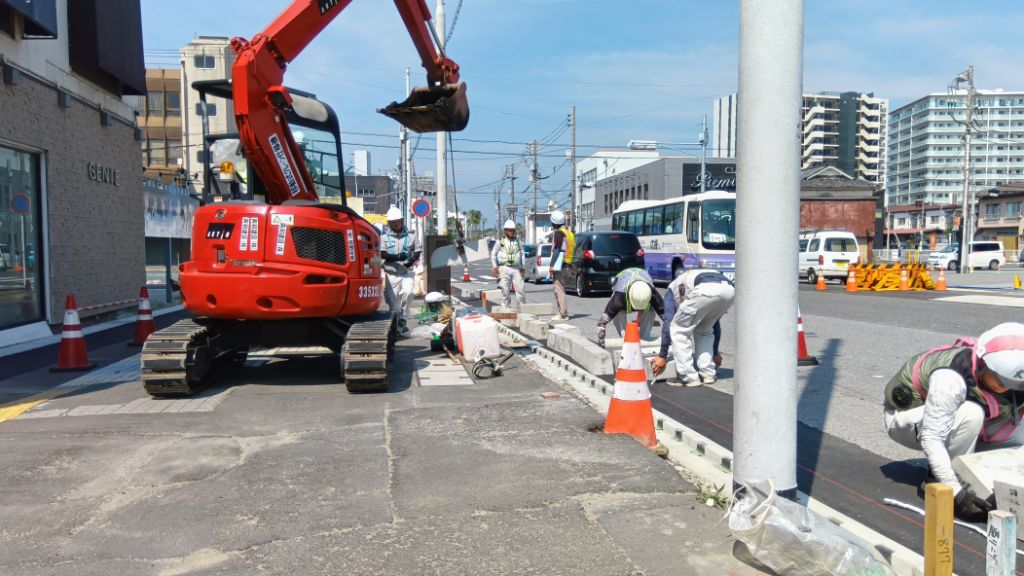 油圧ショベルで吊り下げた歩車道境界ブロックを数名の作業員で設置していく様子
