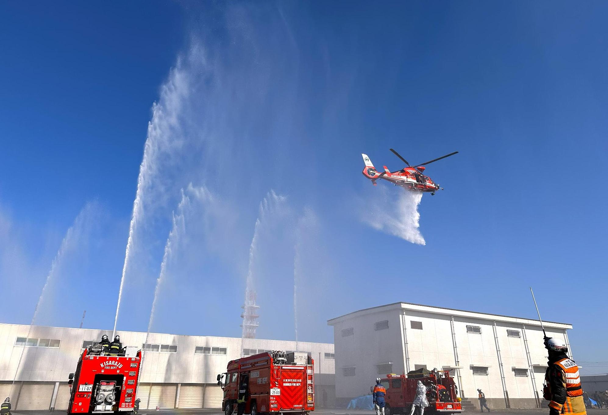 消防車3台と消防士による上空へ放水している様子と、上空よりヘリコプターによる放水をしている様子。