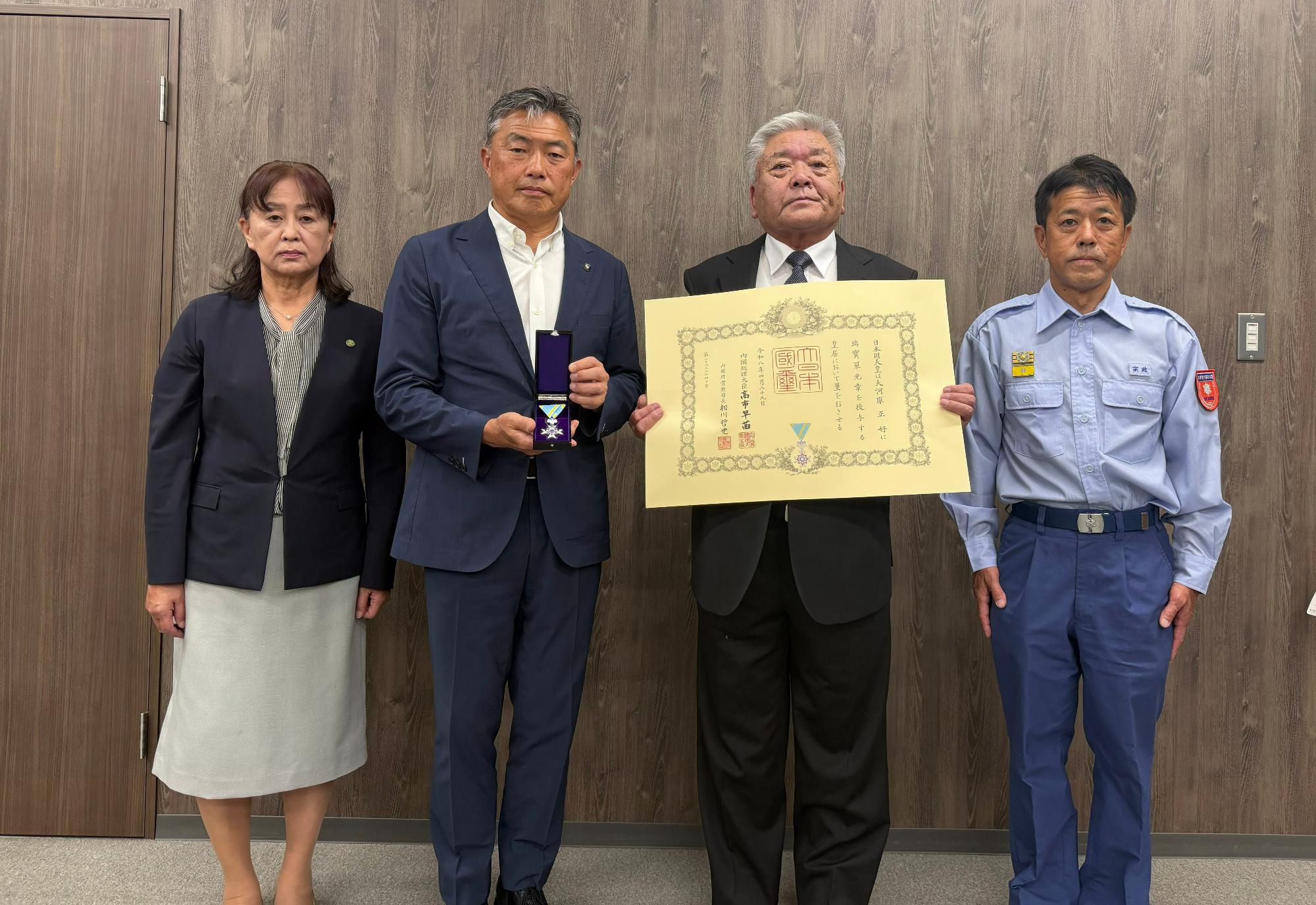 勲章を持った市長と勲記を両手で持った大河原氏が中央に立ち、左側にスーツ姿の女性、右側に制服姿の男性職員が並んで立っている記念写真