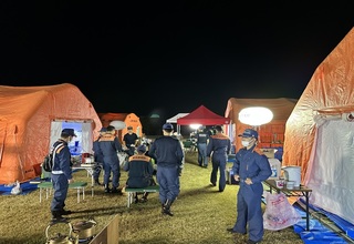太陽が沈み照明が照らされた屋外で、左右に2つずつオレンジ色のテントが設置された間に、隊員の方々が集まっている様子の写真