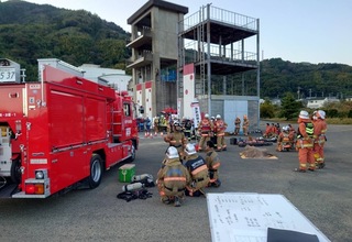 訓練塔の手前に消防車が停車し、周りでヘルメットに防火服を着用した団員の方々が集まっている訓練の様子の写真