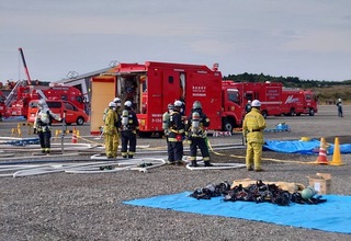 沢山の消防車が停まっている広場に、水色のビニールシートが敷かれ、ホースを伸ばした場所に防火服を着用した団員の方々が立っている訓練の様子の写真