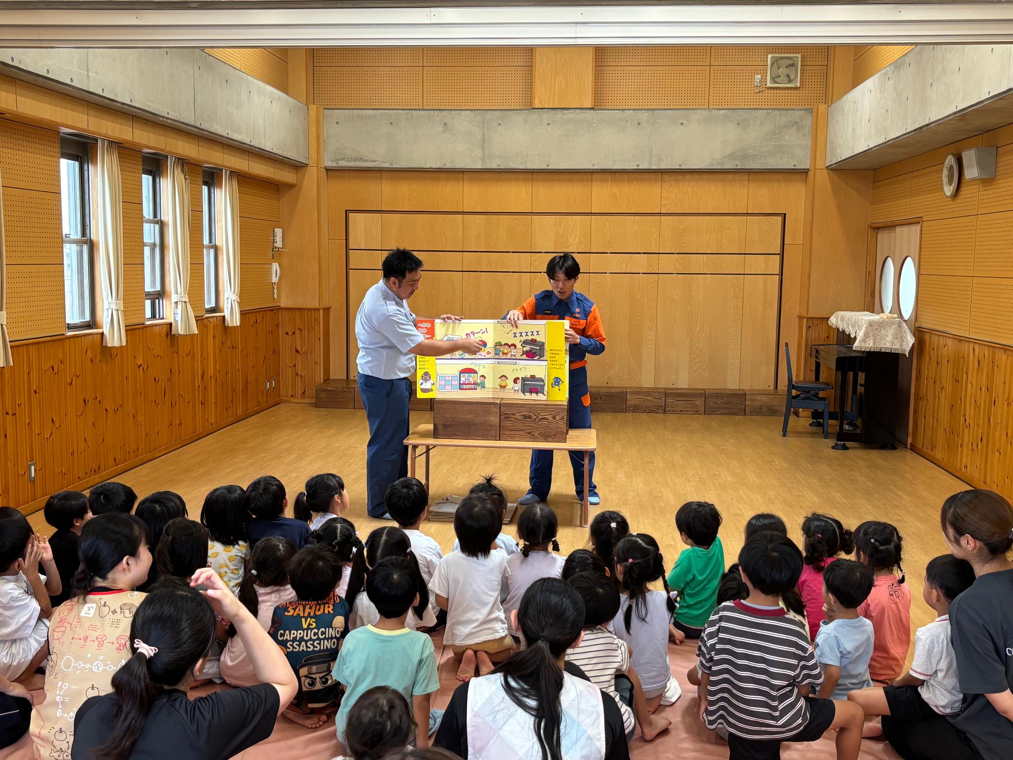 請西保育園内の部屋で消防団学生部1名と消防本部警防課員1名が座っている大勢の園児の前で防災啓発に関する紙芝居をしている様子