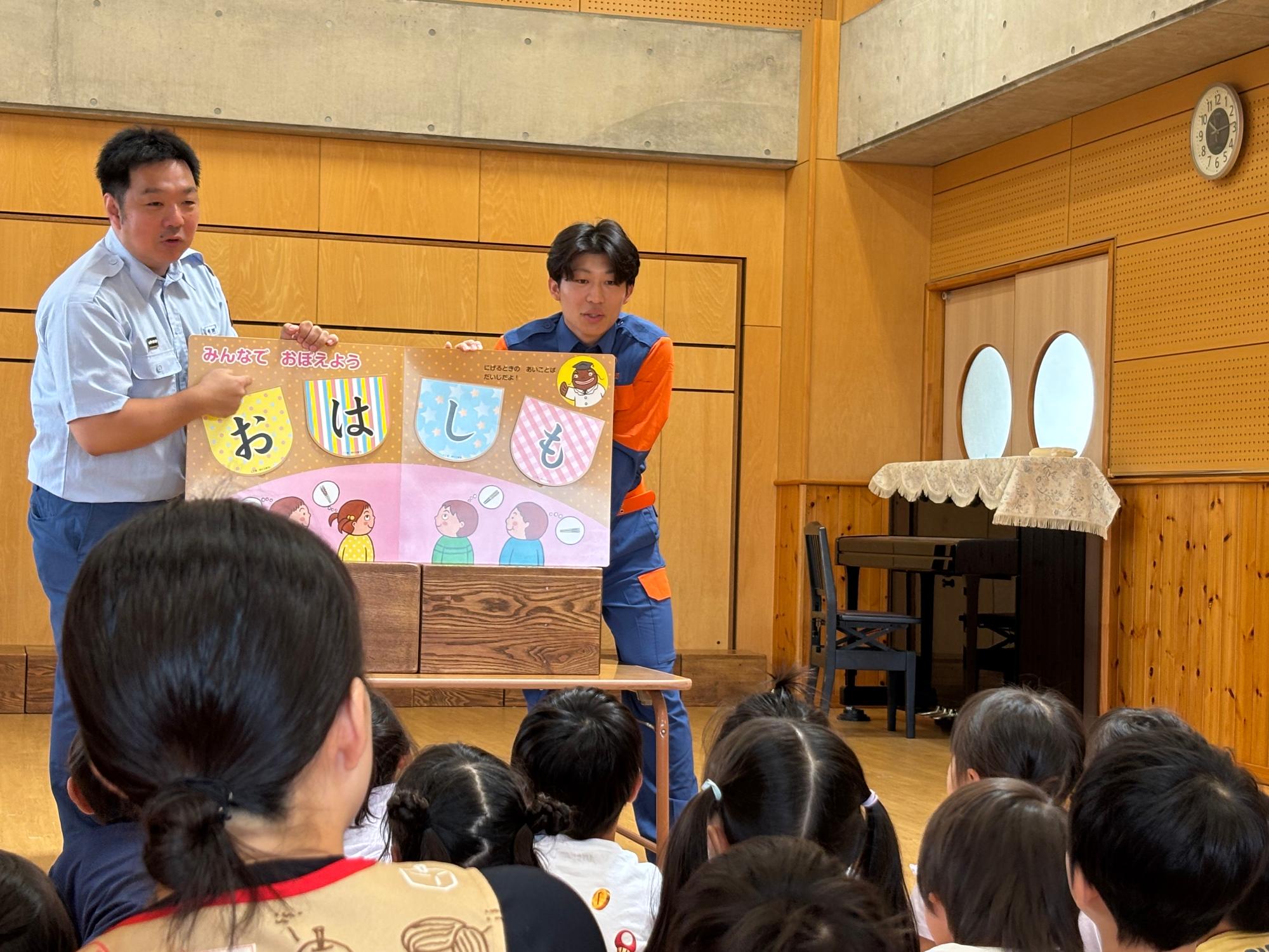 請西保育園内の部屋で消防団学生部1名と消防本部警防課員1名が座っている大勢の園児の前で防災啓発に関する紙芝居をしている様子