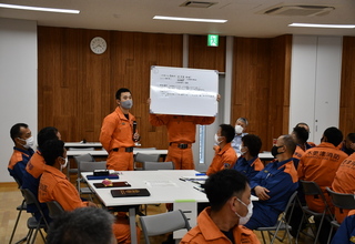 グループごとに分かれて座っている隊員の方々が、前方で1名の隊員が小さなホワイトボードを持っている左側で、もう一人の隊員が図上訓練の結果を発表している様子の写真