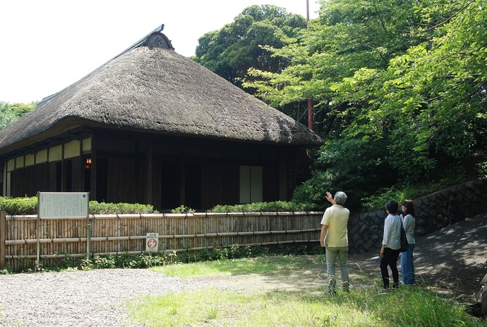 竹垣の中に建っている藁ぶき屋根の旧安西家住宅を見ながら、竹垣の外に立ったガイドの男性が、隣の女性2人に解説をしている様子の写真