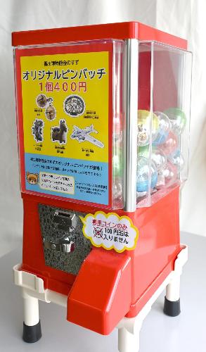 赤いカプセルトイのマシンのカラー写真。マシンの中にはピンバッチを入れた色とりどりのカプセルがたくさん見えます。