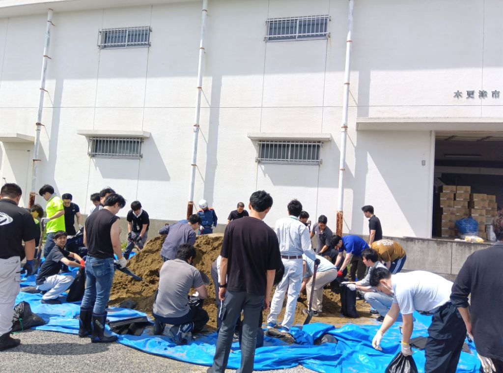 ブルシートの上にある大量の土を大人数の職員がスコップを使用して、土のう袋に入れて土のうを作成している様子です。