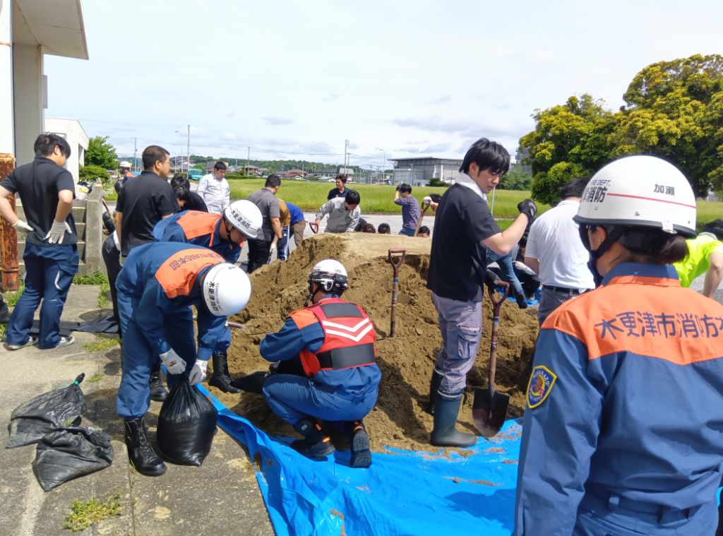 ブルシートの上にある大量の土を大人数の職員がスコップを使用して、土のう袋に入れて土のうを作成している様子です。