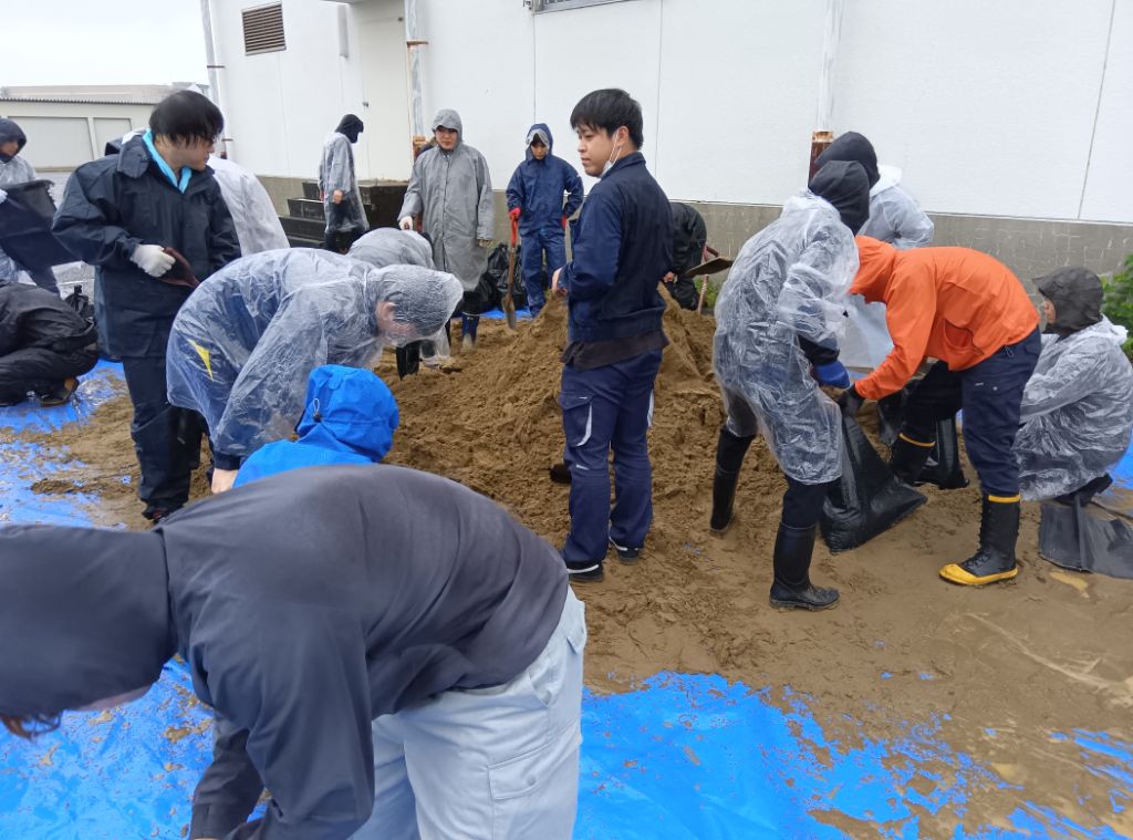 ブルシートの上にある大量の土を大人数の職員がスコップを使用して、土のう袋に入れて土のうを作成している様子です。