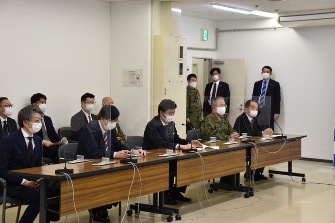 アクリル板で仕切られた長机に5名の関係者が座り、後方の椅子に4名、奥側の壁に3名の関係者が立っている面会の様子の写真