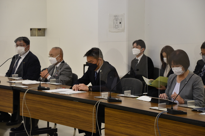 アクリル板で仕切られた長机に4名の関係者が座り、机上の資料を見ている写真
