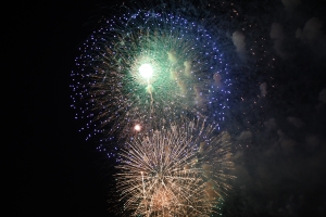 緑・青・白などの色鮮やかな花火と、下には金色の花火が重なる花火大会の一瞬を捉えた写真