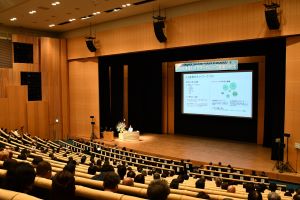 広いホールに参加者が座り、前方のステージでスクリーンを用いて講演が行われている写真