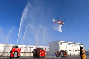 消防車が一斉放水を行い、その上空を消防ヘリコプターが低空で飛行している様子を写した写真