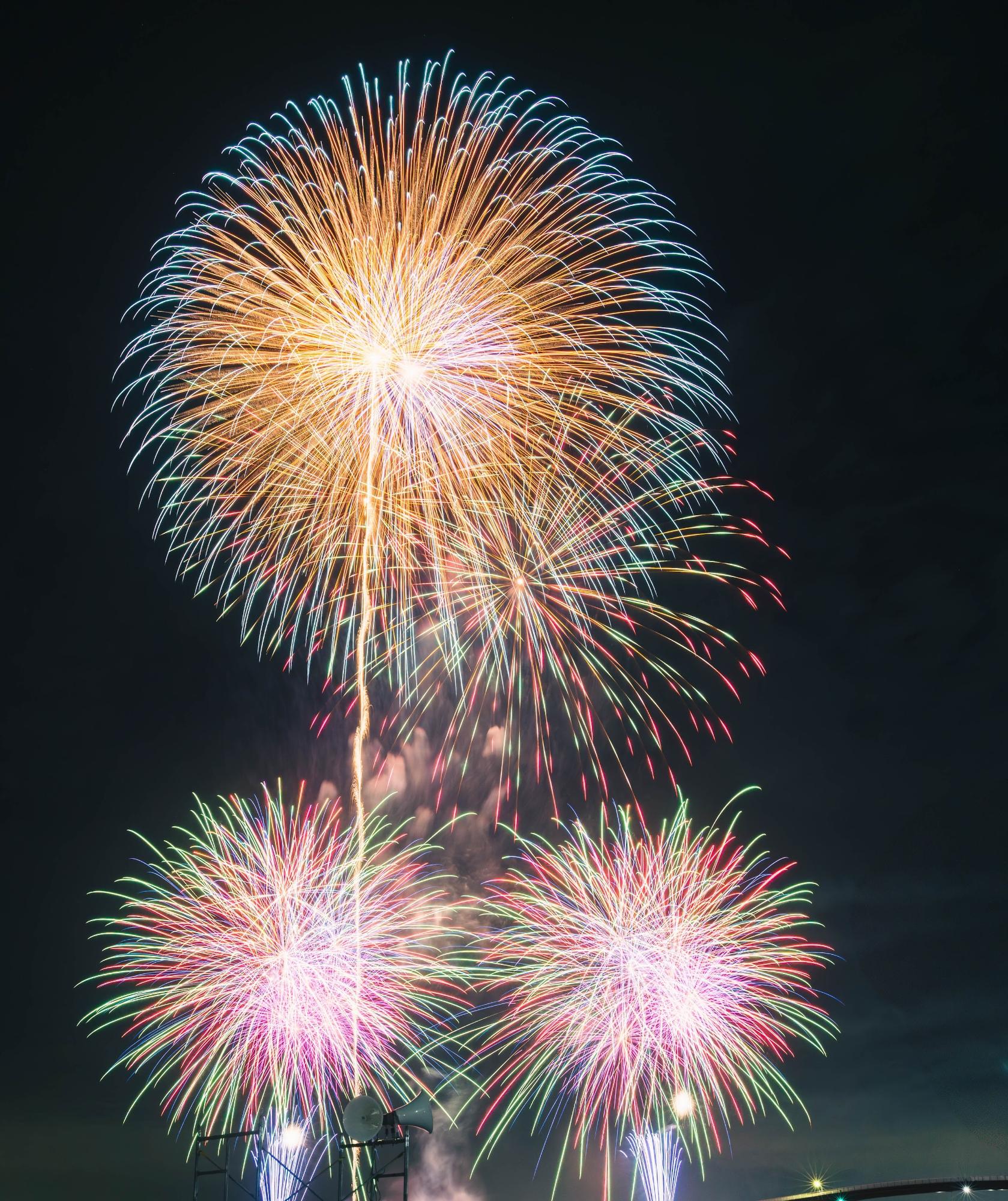 海上に打ちあがった大輪の花火と、その下の3対の打ち上げ花火、噴出花火が色とりどりの光で夜空彩る花火大会の写真