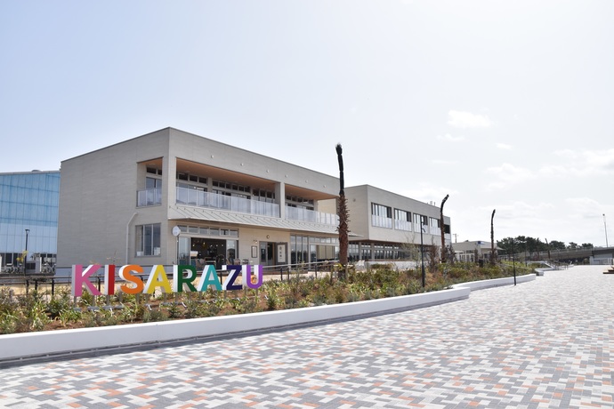植え込みに設置された、1文字ずつ異なる色のアルファベットでKISARAZUとかかれたカラフルなモニュメントと、植え込みの奥にある、白い外壁で2階建ての建物の写真