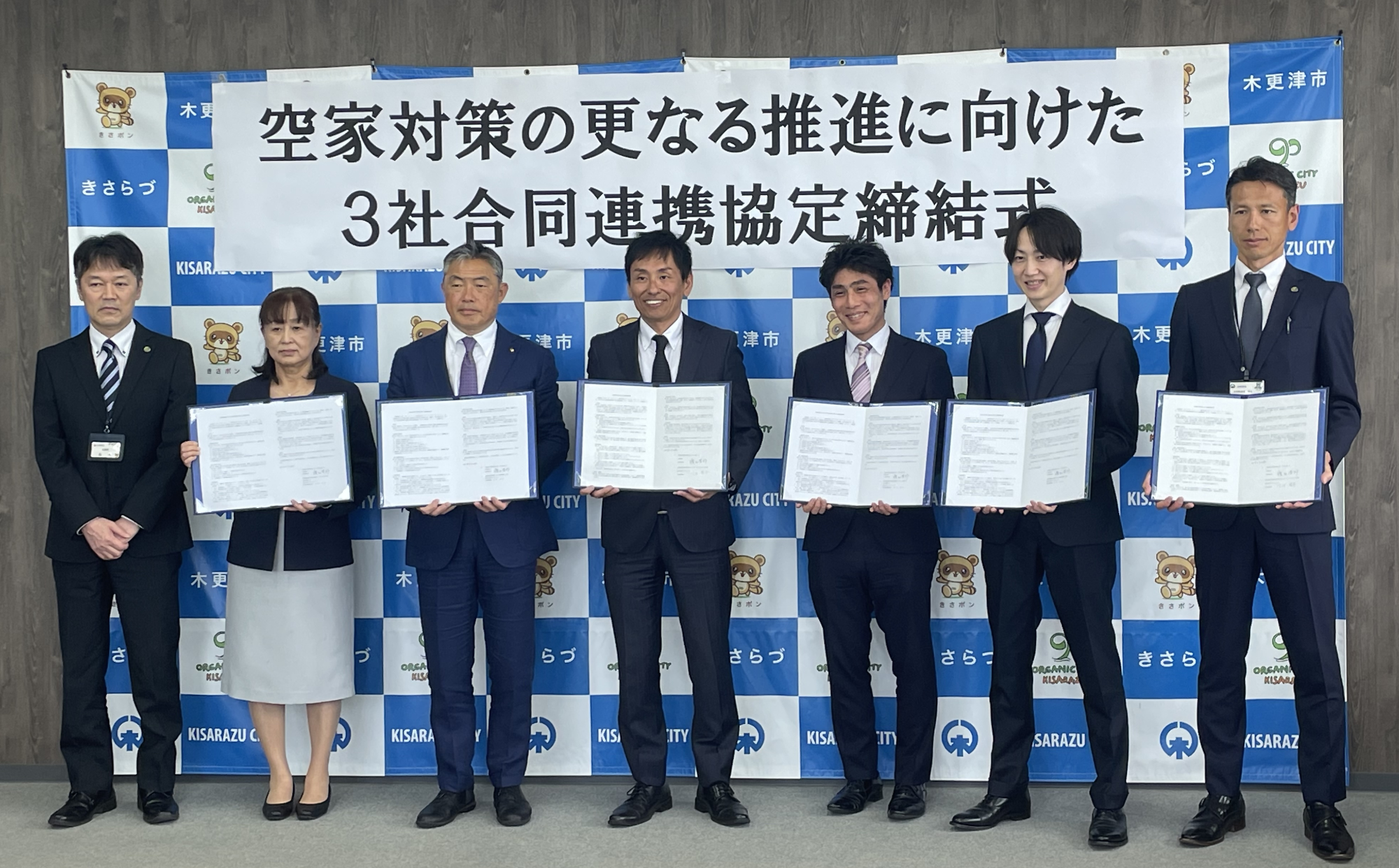 木更津市のバックボードの前で、7名の男女が横一列に並び、協定書を持って記念撮影をしている写真