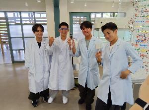 かずさDNA研究所での写真。男子中学生4名が白衣を着て写真に写っている。右後ろにはDNAをモチーフにした模型が写っている。