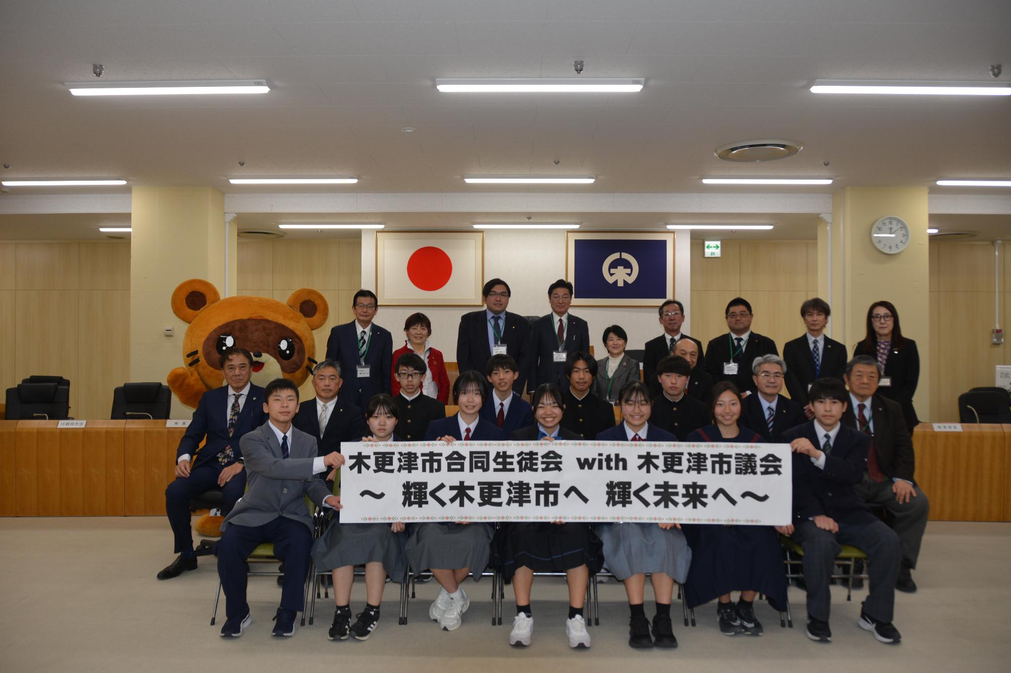 「木更津市合同生徒会with木更津市議会～輝く木更津市へ輝く未来へ～」と書かれた横断幕をもった合同生徒会のメンバー11名と市長、市議会議長、教育長、市議会議員8名、教育委員会事務局が市議会議場前で並んでいる様子の記念写真