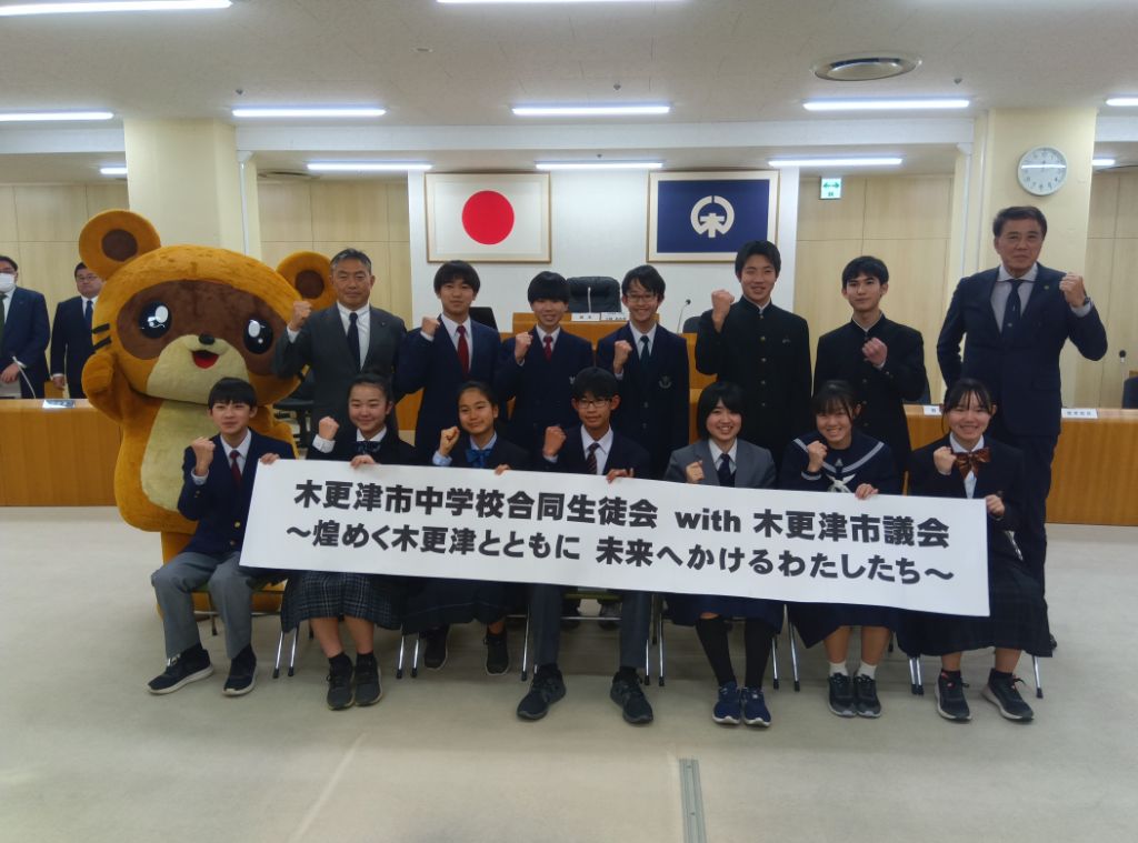 木更津市中学校合同生徒会with木更津市議会煌めく木更津とともに未来へかけるわたちたちと書かれた横断幕をもった合同生徒会のメンバーと12名と市長,教育長,きさポンが市議会議場前で並んでいる様子の記念写真