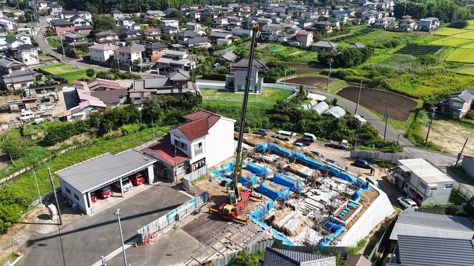 令和7年8月4日時点の消防署富来田分署建設現場を斜め上空から撮影した写真です。写真中央には建設予定地が広がり、建物の基礎工事が進められています。地面にはコンクリート基礎や鉄筋が配置され、青色の養生シートで覆われた掘削部分が複数見られます。現場中央には大型クレーン車が設置され、基礎工事に必要な資材の搬入や組立作業が行われています。左側には既存の消防署庁舎と消防車庫があり、車庫内には消防車両が駐車しています。工事区域は仮囲いで区画され、周辺の住宅地や農地に配慮しながら施工が進められている様子が分かります。この時点では建物本体の着工初期段階であり、建物を支える基礎や柱の土台部分の施工が進んでいる状況です。