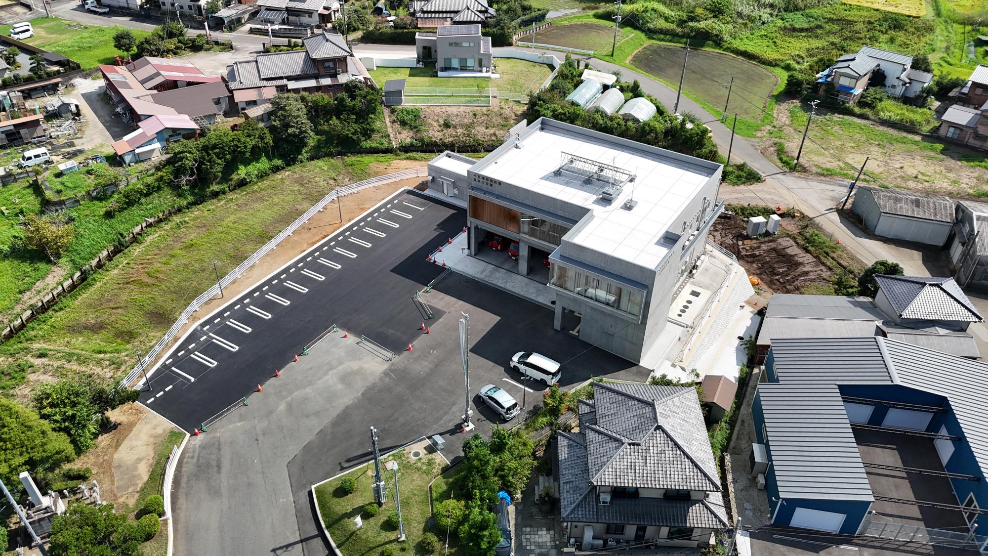 上空から見た消防署富来田分署の建物で、白とグレーを基調とした現代的な2階建ての建物です。建物の正面には消防車などが出入りする大きな車庫があり、その前に黒く舗装された広い駐車場があります。駐車場には白線で区切られた駐車スペースが多数あり、白い車やシルバーの車が数台停まっています。建物の周囲には工事用のカラーコーンや柵が一部残っています。建物の右側と手前には瓦屋根の住宅があり、奥には田畑やビニールハウス、住宅が広がっています。