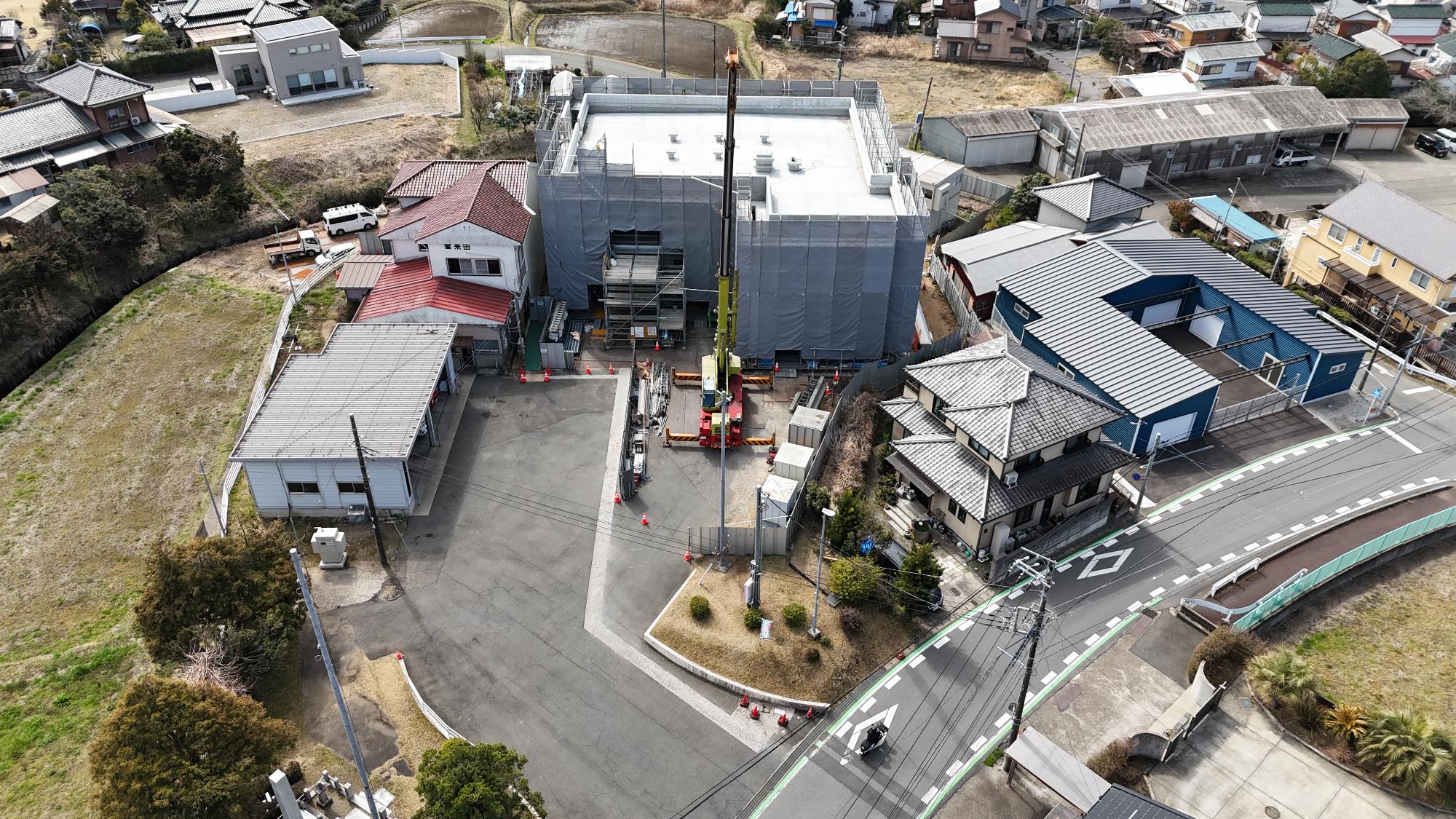 令和8年3月11日時点の消防署富来田分署建設現場を斜め上空から撮影した写真です。写真中央には建設中の新しい分署庁舎があり、建物全体が足場と灰色の養生シートで覆われています。屋上のコンクリート面はすでに完成しており、平らな形状を確認できます。建物の正面には大型クレーン車が配置され、資材搬入や設備設置の作業が行われている様子です。工事区域の出入口や周囲には多数のカラーコーンが設置され、安全対策が講じられています。建設中の建物の左側には、赤い屋根を持つ既存の消防施設があり、その建物には「富来田」と読める表示が見えます。右側には青色の外壁を持つ大きな倉庫や車庫があり、その前には道路が通っています。周辺には住宅や農地、空き地が広がっており、新しい分署庁舎が地域の生活環境に隣接して整備されている状況が分かります。この写真からは、建物の骨組みや屋根の工事がほぼ完了し、外壁や設備工事など仕上げ段階に向けて工事が進んでいる様子がうかがえます。
