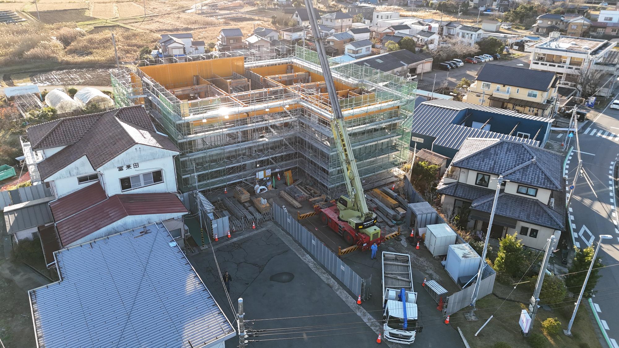 令和7年12月15日時点の消防署富来田分署建設現場を斜め上空から撮影した写真です。写真中央には建設中の新庁舎があり、建物全体が足場と緑色の安全ネットで囲まれています。建物の最上部では黄色い型枠が立ち上がり、部屋ごとの区画が分かる状態となっています。まだ屋根や上階床の施工途中で、建物内部の間取りを確認できる段階です。建物正面の工事ヤードには大型クレーン車が配置され、木材や鉄筋、型枠材などの資材が整理して置かれています。建設中の建物の左側には既存の消防署庁舎があり、「富来田」と書かれた建物と消防車庫が見えます。周辺には住宅地や農地が広がっており、地域に囲まれた環境の中で工事が進められている様子が分かります。この時点では建物の躯体工事が進行中で、上階の壁や床を構築する重要な工程にあることが確認できます