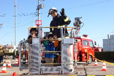 はしご車搭乗体験をしている子ども2人が消防隊員と一緒にカメラに向かって手を振っている様子