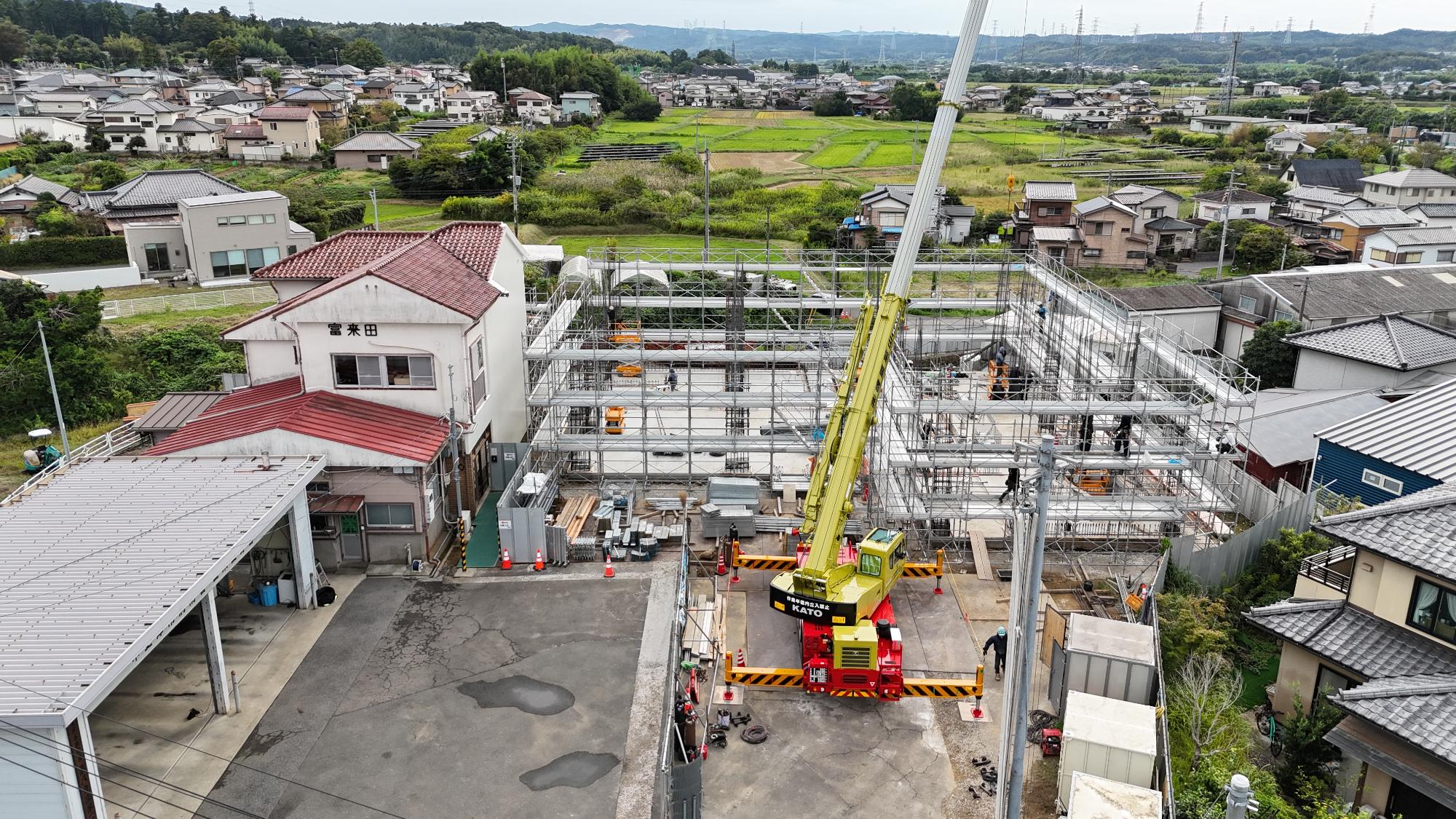 令和7年10月3日時点の消防署富来田分署建設現場を斜め上空から撮影した写真です。写真中央には建設中の新庁舎があり、足場に囲まれた建物内部では柱や梁となる鉄筋、型枠の組立作業が進められています。建物前面には大型クレーン車が配置され、資材搬入や鉄骨・型枠材の揚重作業が行われています。左側には既存の消防署庁舎があり、「富来田」の表示が確認できます。周辺には住宅地や田畑が広がり、地域に囲まれた環境の中で工事が進められている様子が分かります。この時点では建物の土台と1階部分の躯体工事が進行中であり、今後の上階施工に向けて建物の骨格を形成している重要な工程であることが確認できます。