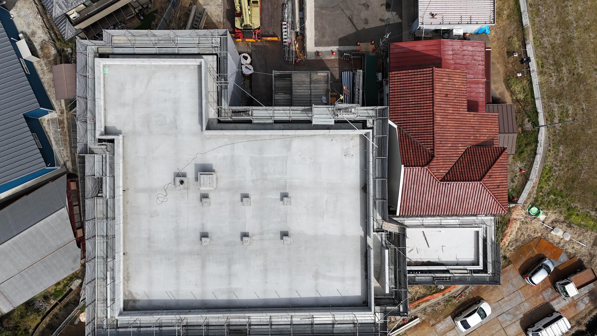 令和8年3月11日時点の消防署富来田分署建設現場を真上から撮影した写真です。建物はほぼ正方形に近い大きな形をしており、屋上のコンクリート床が完成しています。屋上の周囲には転落防止のための足場と養生シートが設置されています。表面は明るい灰色のコンクリートで、施工を終えたばかりのような滑らかな状態です。建物の東側には既存の赤茶色の屋根を持つ旧庁舎建物が隣接しており、その周囲にも工事用の足場が見えます。さらに建物の周辺には住宅や空き地、駐車中の車両があり、地域の住宅地の中で建設工事が進められている様子が確認できます。この写真からは、建物の主要な躯体工事が完了し、屋上部分まで形が整った段階であることが分かります。