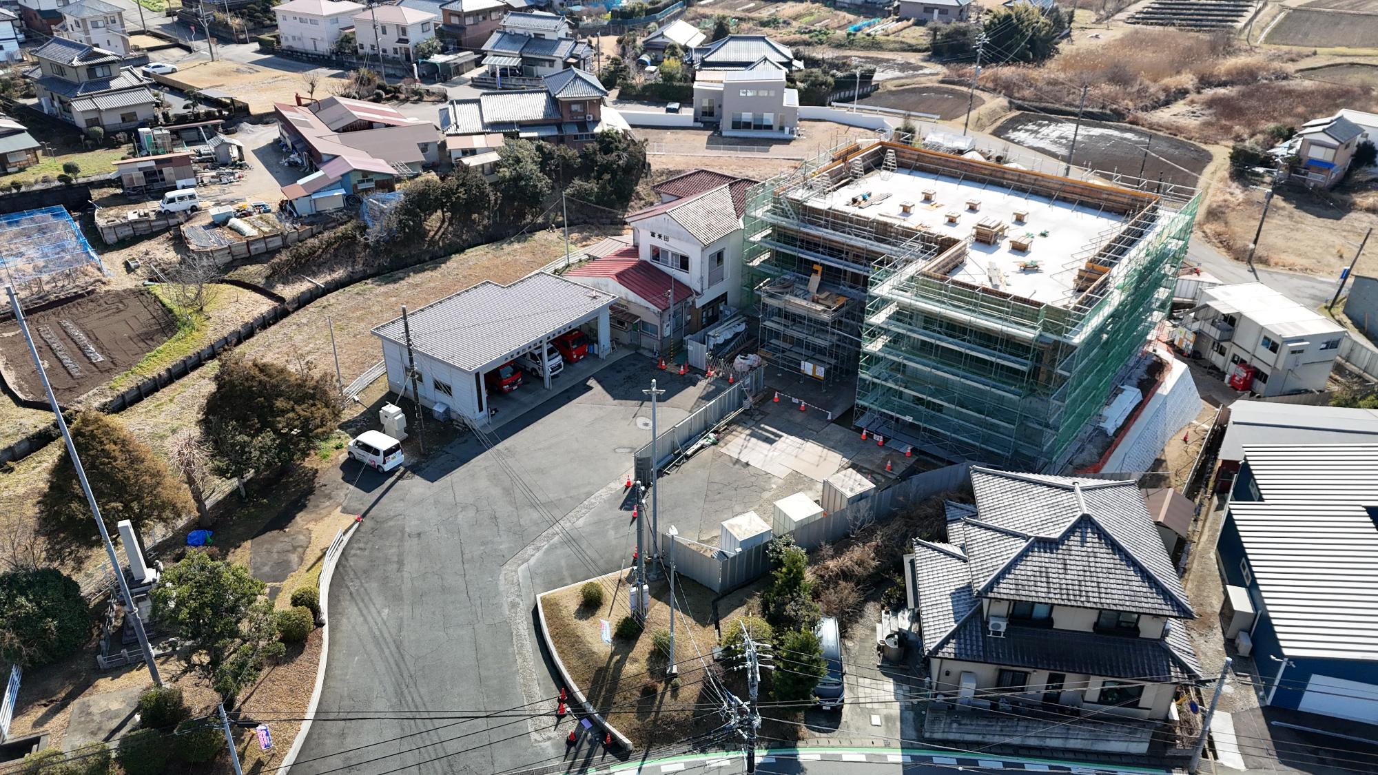 令和8年2月12日時点の消防署富来田分署建設現場を斜め上空から撮影した写真です。写真中央には建設中の新しい分署庁舎があり、建物全体が足場と緑色の安全ネットで覆われています。屋上は平らなコンクリート造で、設備設置用の基礎や資材が並んでいます。建設現場の手前には工事用仮囲いが設けられ、その内部には資材や仮設設備が配置されています。建物の正面には既存の消防施設があり、車庫には赤色の消防車両が複数駐車している様子が確認できます。建物には「富来田」の表示が見えます。周辺には住宅や農地が広がり、地域の生活環境に囲まれた場所で工事が進められていることが分かります。道路沿いや工事区域には多数のカラーコーンが配置され、安全確保に配慮した施工が行われています。この写真からは、建物の主要構造が完成し、足場を残した状態で外壁工事や内部設備工事、仕上げ工事が進められている段階であることがうかがえます。なお、令和8年3月の写真と比較すると、屋上設備や周辺施設の整備が進む前の状況であり、建設工事が着実に進捗している様子が確認できます。