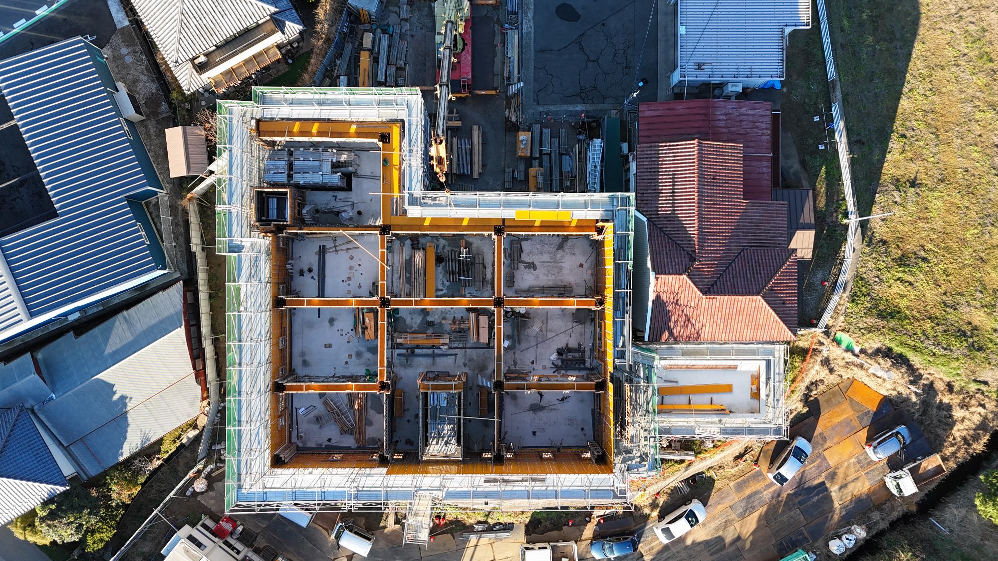 令和7年12月15日時点の消防署富来田分署建設現場を真上から撮影した写真です。建設中の建物はほぼ四角形の形状をしており、周囲を足場が囲んでいます。建物内部では黄色い型枠によって複数の部屋や廊下が区切られており、将来の庁舎内部の配置が見て取れます。床部分はコンクリートで施工されている一方、上階床や屋根にあたる部分はまだ完成しておらず、鉄筋や型枠材が設置された状態です。建物の右側には赤茶色の屋根を持つ既存施設が隣接し、その周囲でも工事用足場が設けられています。敷地内には建設資材や作業用車両が配置され、資材搬入や組立作業が継続して行われている様子が確認できます。この写真からは、建物の骨組みや内部の構造が形成され、屋上スラブ施工前の段階まで工事が進んでいることが分かります。