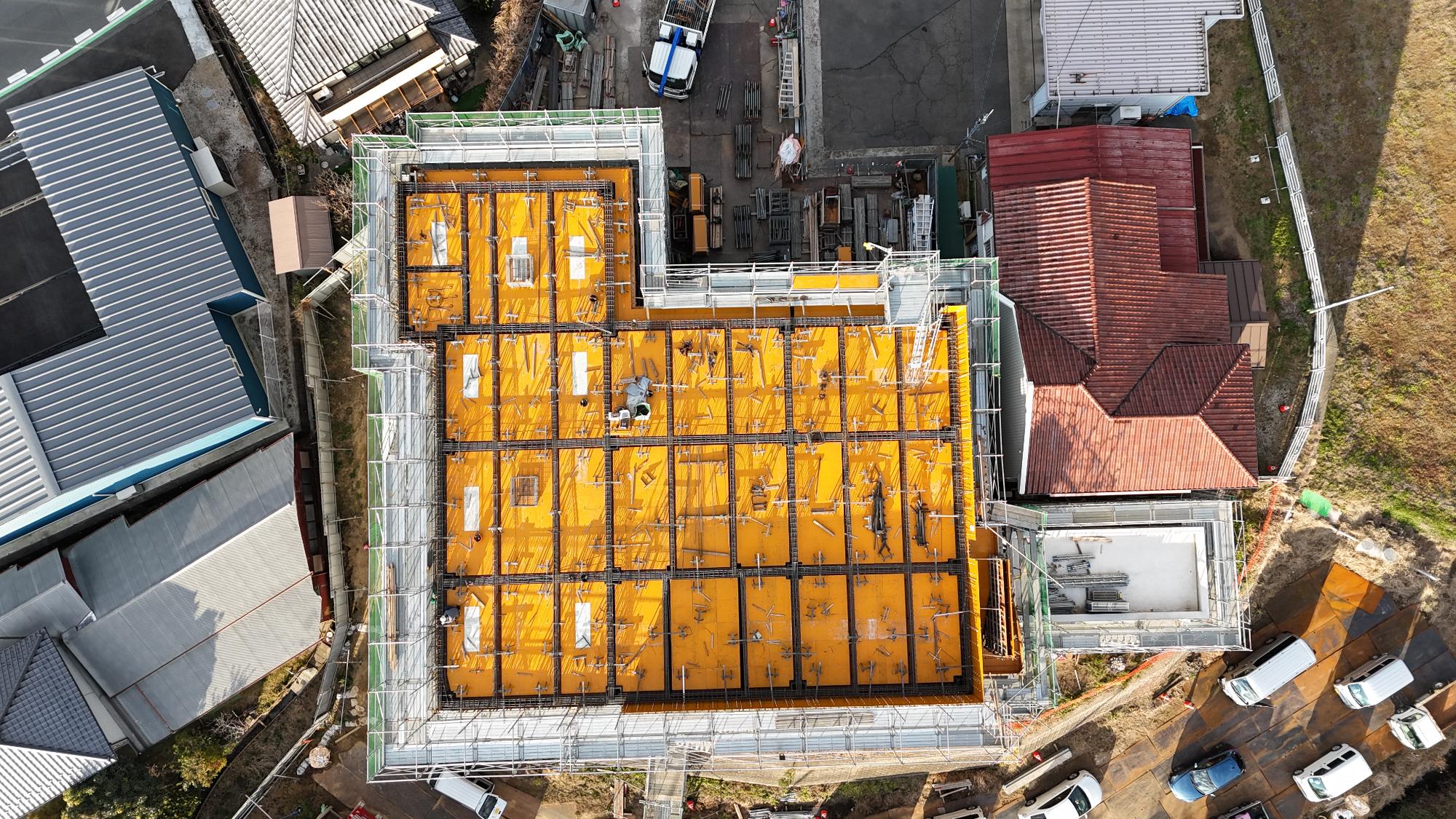 令和8年1月9日時点の消防署富来田分署建設現場を真上から撮影した写真です。建設中の建物は大きな四角形で、建物全体を囲むように足場と安全ネットが設置されています。屋上部分はまだ完成しておらず、鮮やかな黄色の型枠が広い面積を覆っています。型枠の上には縦横に格子状の鉄筋が組まれ、コンクリートを打設する前の状態となっています。ところどころに設備用の開口部や突起が見られ、複数の作業員が屋上で施工を行っています。建物の東側には赤茶色の屋根を持つ既存の消防施設が隣接しており、その南側には増築部分と思われる小規模な構造物も確認できます。現場周辺には資材置場や駐車中の車両が配置され、工事が活発に進められている様子がうかがえます。この写真からは、建物本体の骨組みと上階部分の施工が進み、屋上スラブのコンクリート打設を控えた段階であることが分かります。