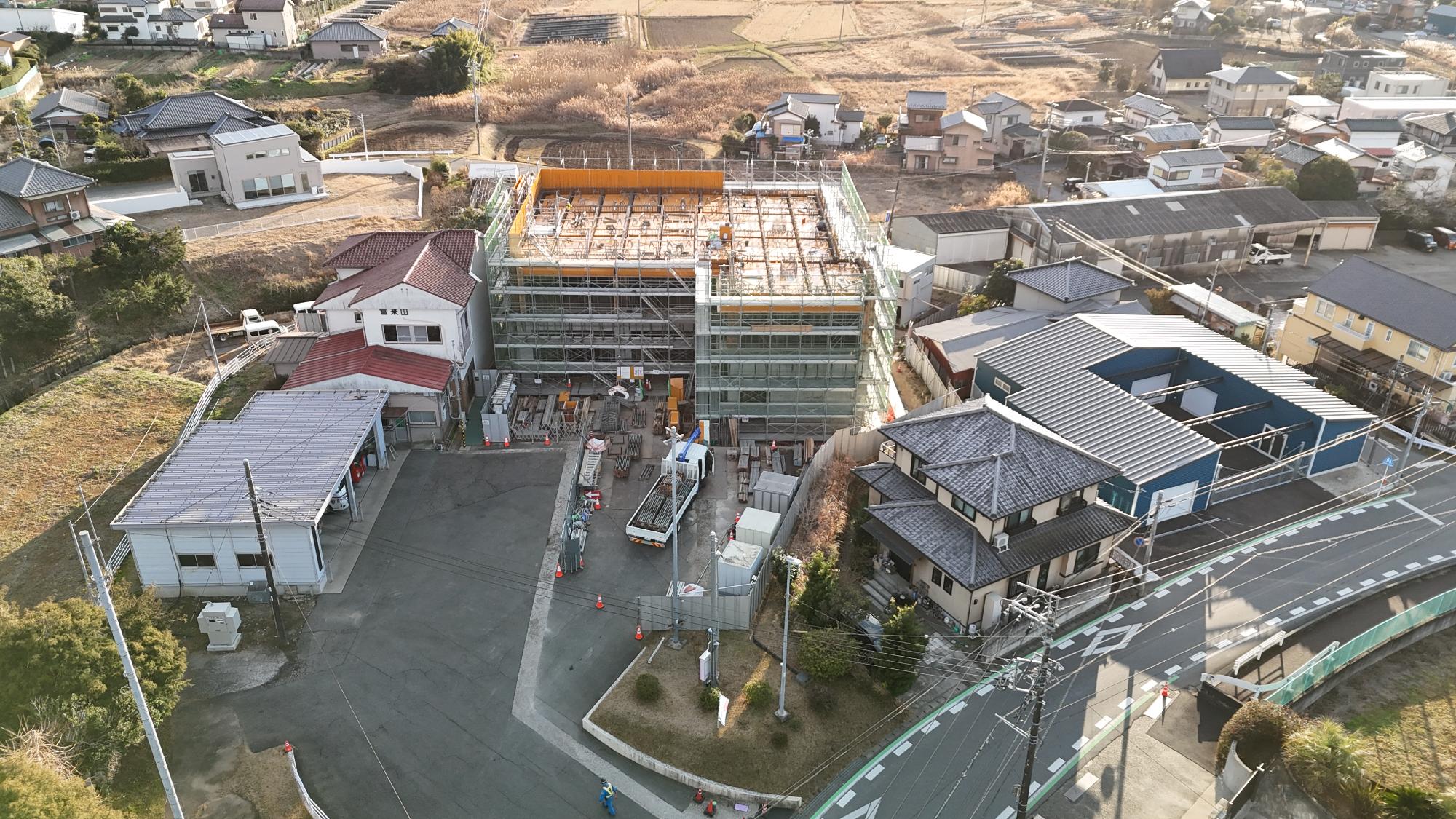 令和8年1月9日時点の消防署富来田分署建設現場を斜め上空から撮影した写真です。写真中央に建設中の新しい分署庁舎があり、建物全体が足場と緑色の安全ネットで覆われています。最上部には明るい黄色の型枠と鉄筋が広がり、屋上階の施工が進められている状況です。建設中の建物の左側には既存の消防署庁舎があり、建物正面には「富来田」の文字が確認できます。その手前には消防車庫があり、赤色の消防車両が格納されています。建物正面の工事ヤードにはトラックや建設資材が集積され、搬入や組立作業が行われています。工事区域の周囲には柵やカラーコーンが設置され、安全管理が徹底されています。周辺には住宅地や農地が広がり、新旧の消防施設が並んで存在している様子が確認できます。この時点では、建物の主要な鉄筋コンクリート躯体工事が終盤を迎え、屋上部分のコンクリート打設に向けた準備が進められている段階です。