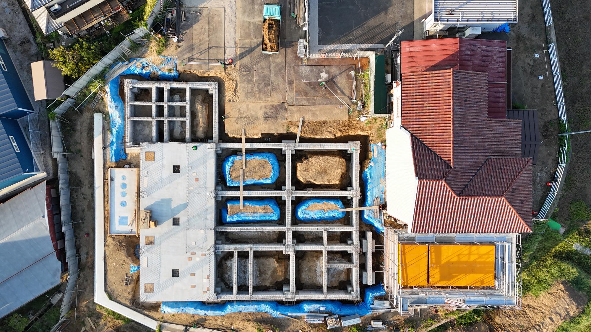 令和7年9月10日時点の消防署富来田分署建設現場を真上から撮影した写真です。建設区域ではコンクリートの基礎梁が碁盤目状に組まれ、その内部に土が残された区画が規則的に並んでいます。一部には青色の養生シートが敷かれ、基礎の保護や施工管理が行われています。建物の左側では白いコンクリート床が施工されており、将来の車庫や附属施設の基礎部分と考えられます。右側には赤茶色の屋根を持つ既存の消防施設が隣接しており、新庁舎の建設が既存施設のすぐ隣で進められていることが分かります。この写真からは、建物の土台となる基礎工事がほぼ形を整え、今後の柱や壁の施工に向けた準備が進んでいる段階であることが確認できます。