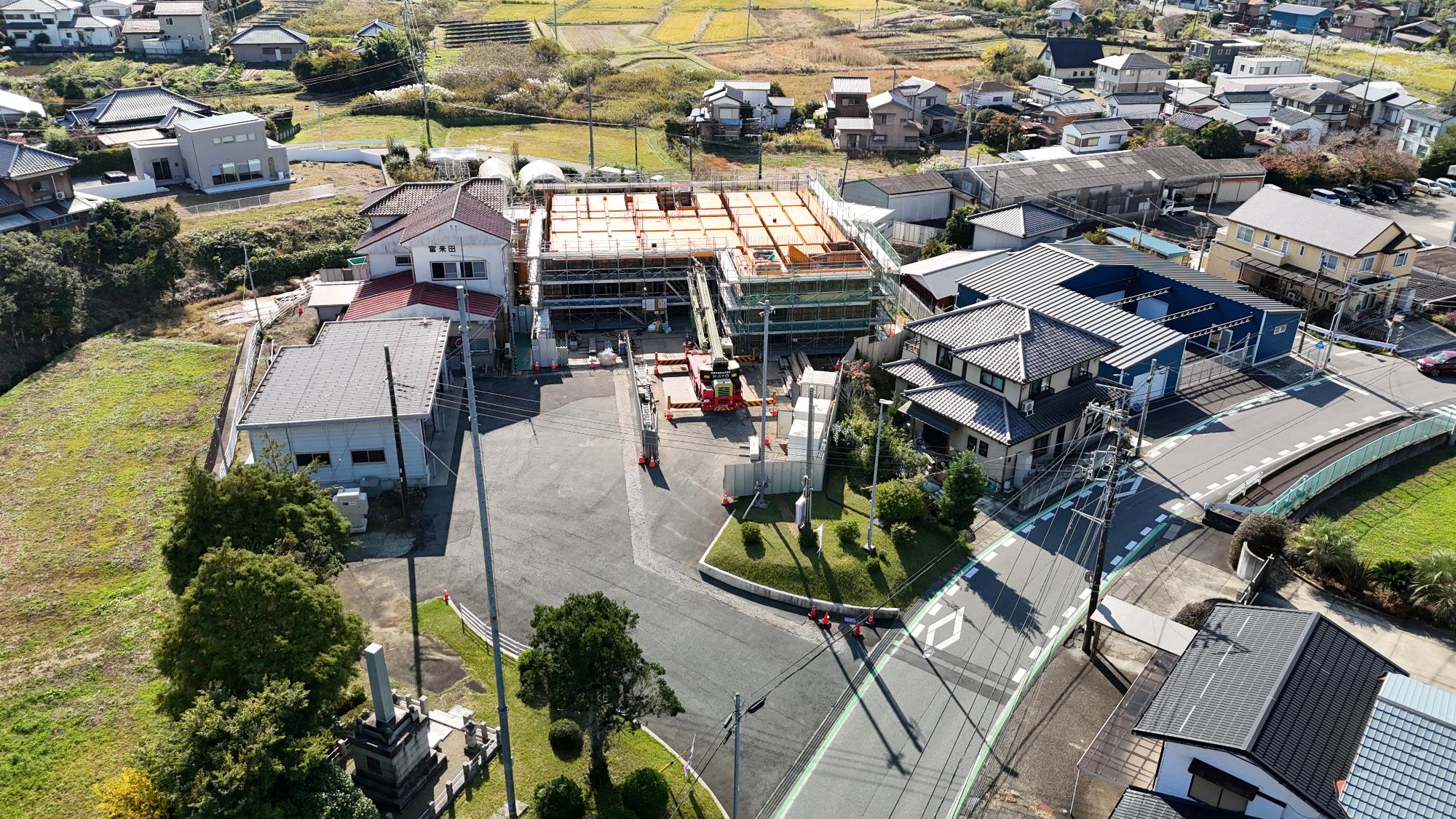 令和7年11月7日時点の消防署富来田分署建設現場を斜め上空から撮影した写真です。写真中央には建設中の新庁舎があり、建物全体を足場と安全ネットが囲っています。建物の上部は鮮やかな黄色の型枠で覆われており、2階部分の床や梁の施工が進められている様子が確認できます。建物正面には大型クレーン車が配置され、資材の搬入や組立作業が行われています。工事区域は仮囲いで区画され、資材や工具類が整然と配置されています。建設中の建物の左側には既存の消防署庁舎と車庫があり、「富来田」の表示が見えます。周辺には住宅地や農地が広がり、地域に密着した環境の中で工事が進められていることが分かります。この時点では建物の主要構造が立ち上がり、上階部分の床や壁を形成する躯体工事が進行している段階です。