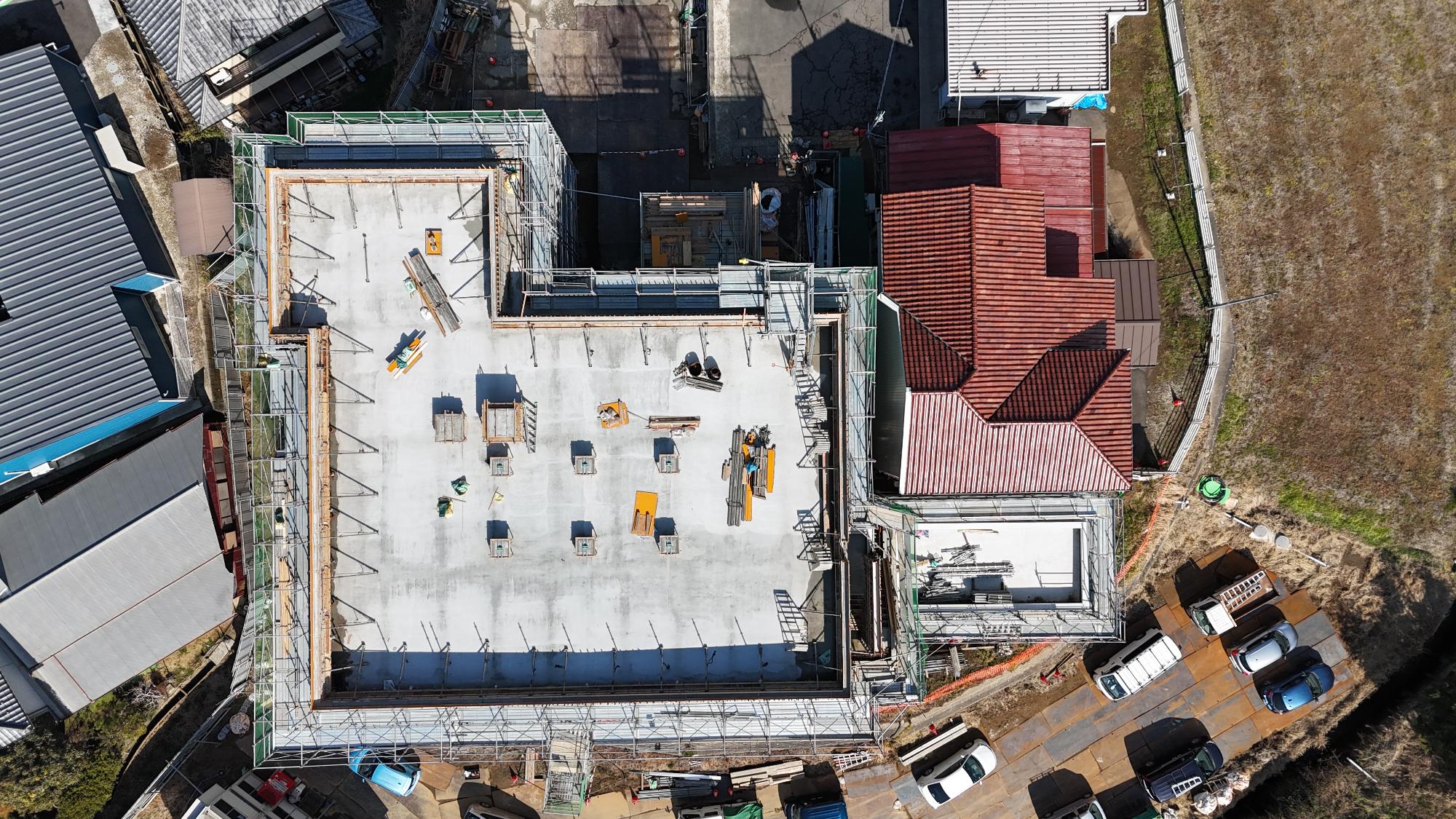 令和8年2月12日時点の消防署富来田分署建設現場を真上から撮影した写真です。建設中の建物は大きな四角形をしており、屋上のコンクリート床が完成しています。建物の周囲には足場が組まれ、安全用のネットや手すりが設置されています。屋上には設備の基礎となる四角い突起が規則的に並んでいるほか、木材や金属製の資材が各所に置かれています。また、屋上の周囲には手すりや設備を取り付けるためと思われる金具が立ち上がっています。建物の右側には赤茶色の屋根を持つ既存の消防施設が隣接しており、その周囲にも工事用の足場が見えます。建物の周辺には工事車両や一般車両が駐車しており、資材置場や作業スペースが確保されています。この時点では建物の躯体と屋上の施工が進み、建物の形がほぼ完成した段階で、今後の外装や設備工事に向けた準備が進められていることが分かります。