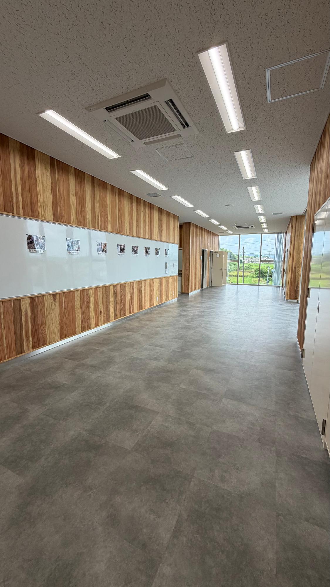 消防署富来田分署の多目的室の写真。消防職員の訓練や、市民向けの救急救命講習を行うための部屋として整備されています。部屋は長方形で、かなり広く感じられます。床には机や椅子などの備品は置かれておらず、大人数が集まったり、訓練用の資機材を広げたりできるように、中央部分が大きく空いています。壁には木目調のパネルが使われており、消防施設にありがちな無機質な印象よりも、温かみのある落ち着いた雰囲気があります。左側の壁には大きなホワイトボードがあり、講習時の説明や図解、訓練内容の共有に利用できます。天井には明るい照明が一直線に並び、室内全体を均一に照らしています。中央には業務用の天井埋込型エアコンが設置されており、多人数が集まる講習会でも快適に過ごせるようになっています。