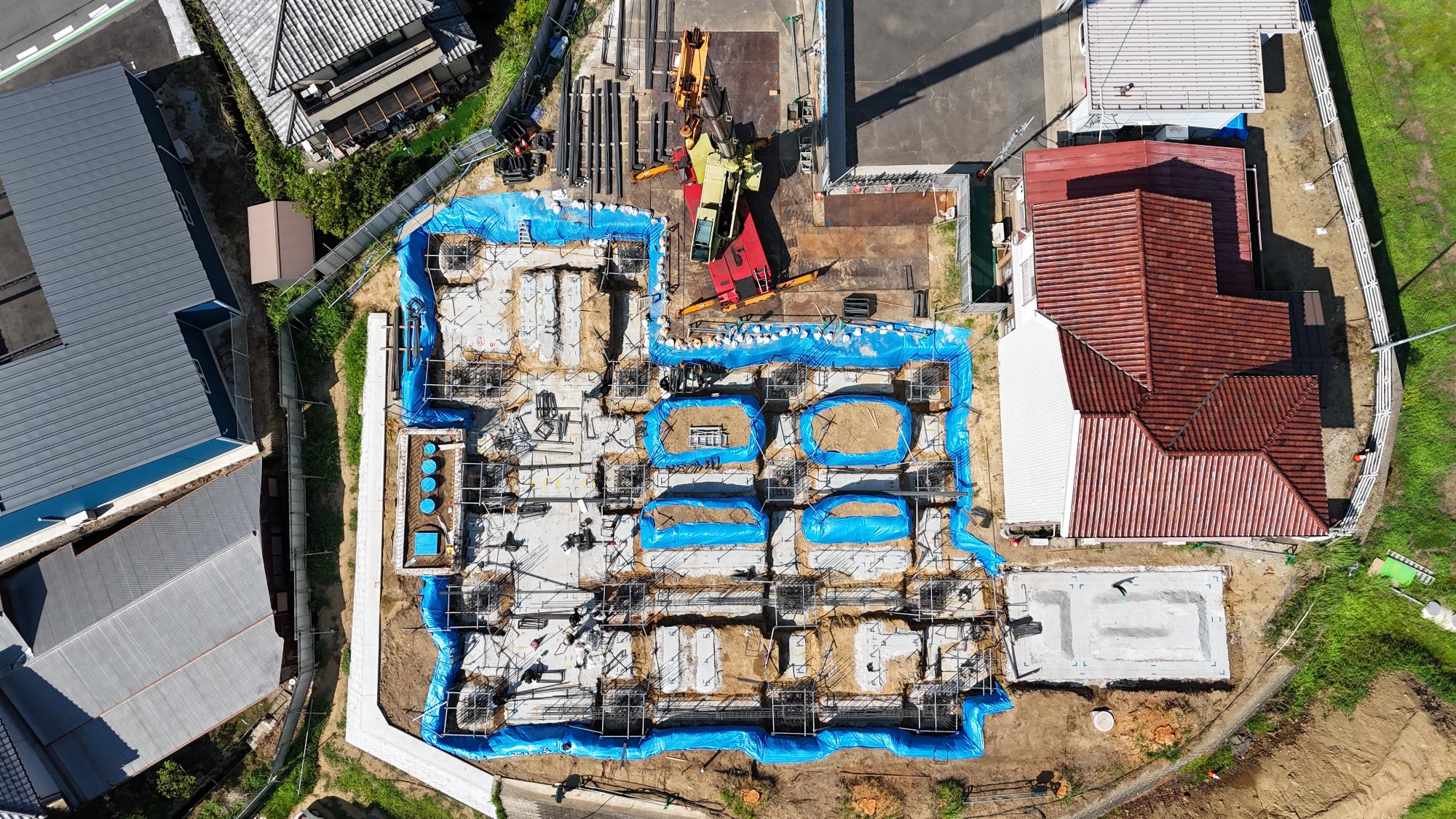 令和7年8月4日時点の消防署富来田分署建設現場を真上から撮影した写真です。建設区域ではコンクリート製の基礎が建物の形に沿って配置され、その上には柱や梁の位置を示す鉄筋が組み立てられています。基礎の周囲や内部には青色の養生シートが敷かれ、雨水や土砂の流入を防ぐ対策が講じられています。建物の輪郭が徐々に見え始めており、将来の部屋や通路の配置を支える基礎構造が形成されています。右側には赤茶色の屋根を持つ既存の消防施設が隣接し、その南側には附属施設の基礎工事も進められています。この写真からは、新庁舎建設の土台となる基礎工事が本格的に進み、建物の骨格づくりに向けた準備が整えられていることが確認できます。