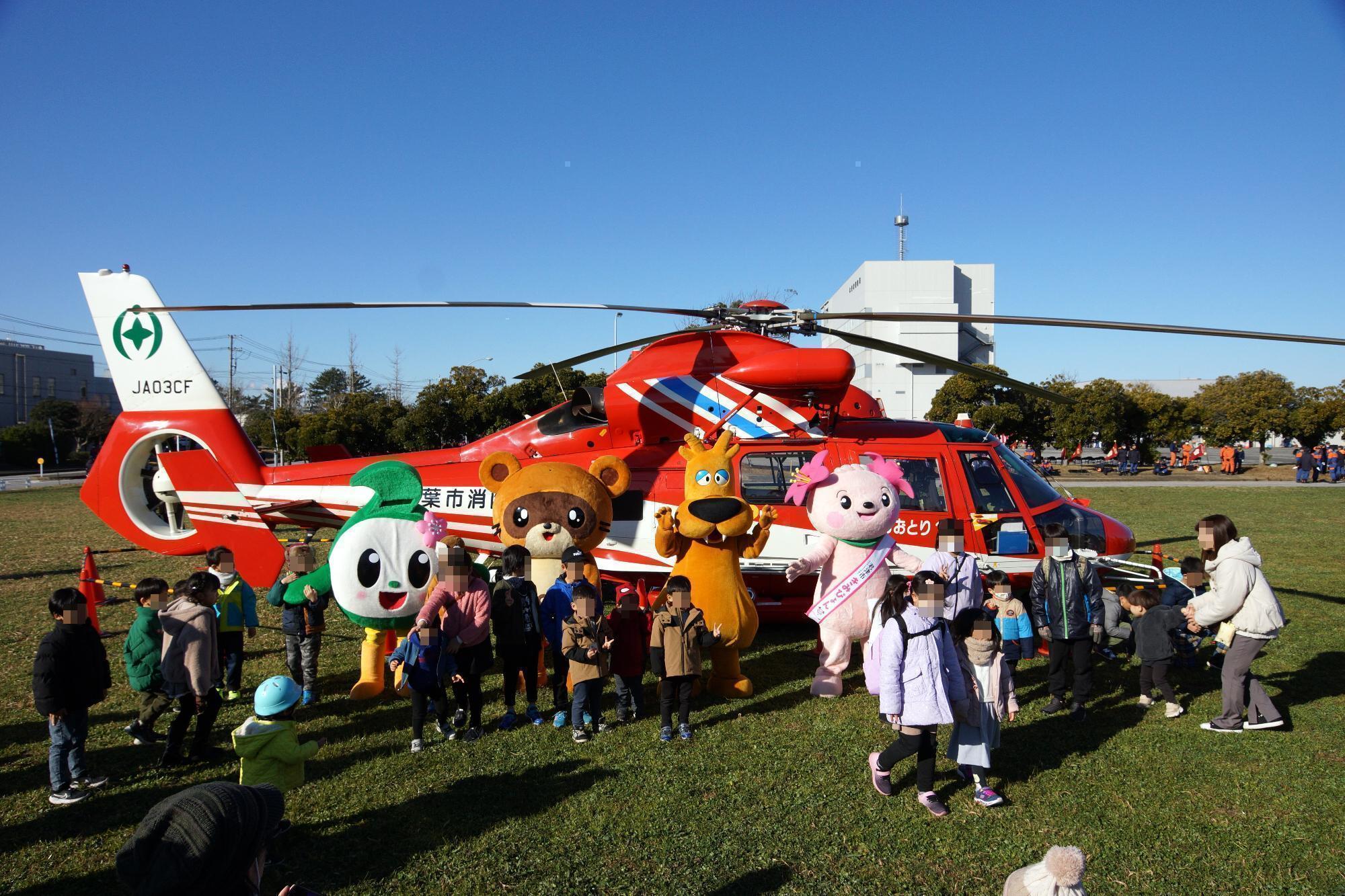 千葉市消防ヘリの前に左からマスコットキャラクターの富津市のふっつん、木更津市のきさぽん、袖ヶ浦市のガウラ、君津市のきみぴょんが立っていて、子どもたちと触れ合っている様子